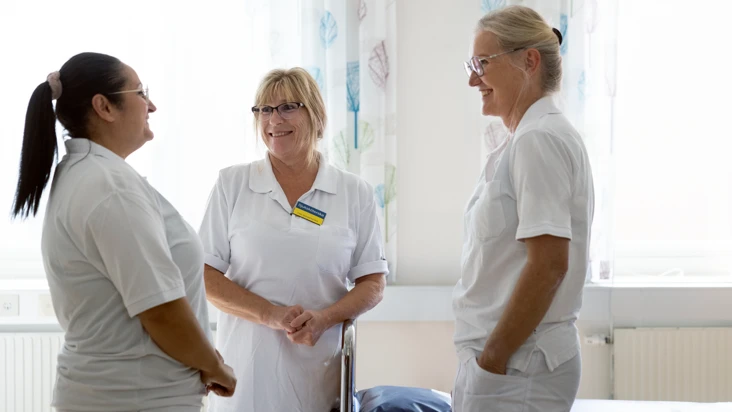tre sjuksköterskor står i ett patientrum på en vårdavdelning och talar gemytligt med varandra.