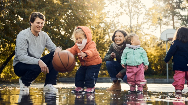 Två förskolebarn leker med basketboll i vattenpöl med två pedagoger bredvid