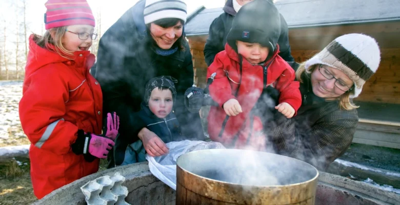 En grupp små barn och vuxna kvinnor som står utomhus runt en kastrull med kokande vatten.
