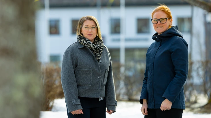 två personer står utomhus i vintermiljö med stort vitt hus i bakgrunden.