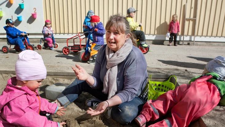 En kvinna sitter i en sandlåda med en grupp förskolebarn.