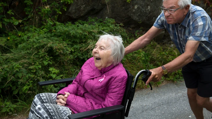 glad äldre kvinna i rullstol blir körd av äldre man som ser lite sammanbiten ut.