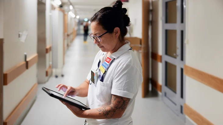 kvinna i vårdkläder, tatuerad på armarna, står i en sjukhuskorridor och tittar i en Ipad.