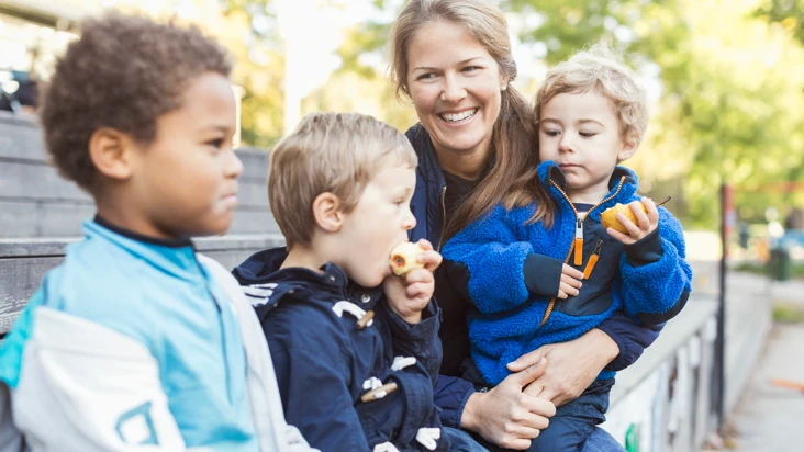 förskollärare tar en paus utomhus i fint sommarväder med tre förskolebarn intill sig.