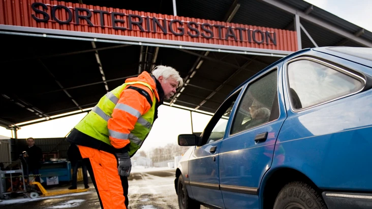 En manlig återvinningsarbetare u orange kläder böjer sig fram mot en bilist i en bil på väg in på återvinningsstationen.
