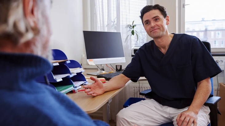 Manlig läkare i blå arbetsblus sitter med öppen handflata och ler mot en patient mitt emot.