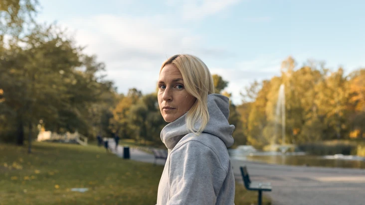 En kvinna i grå huvtröja går utomhus och tittar in i kameran.
