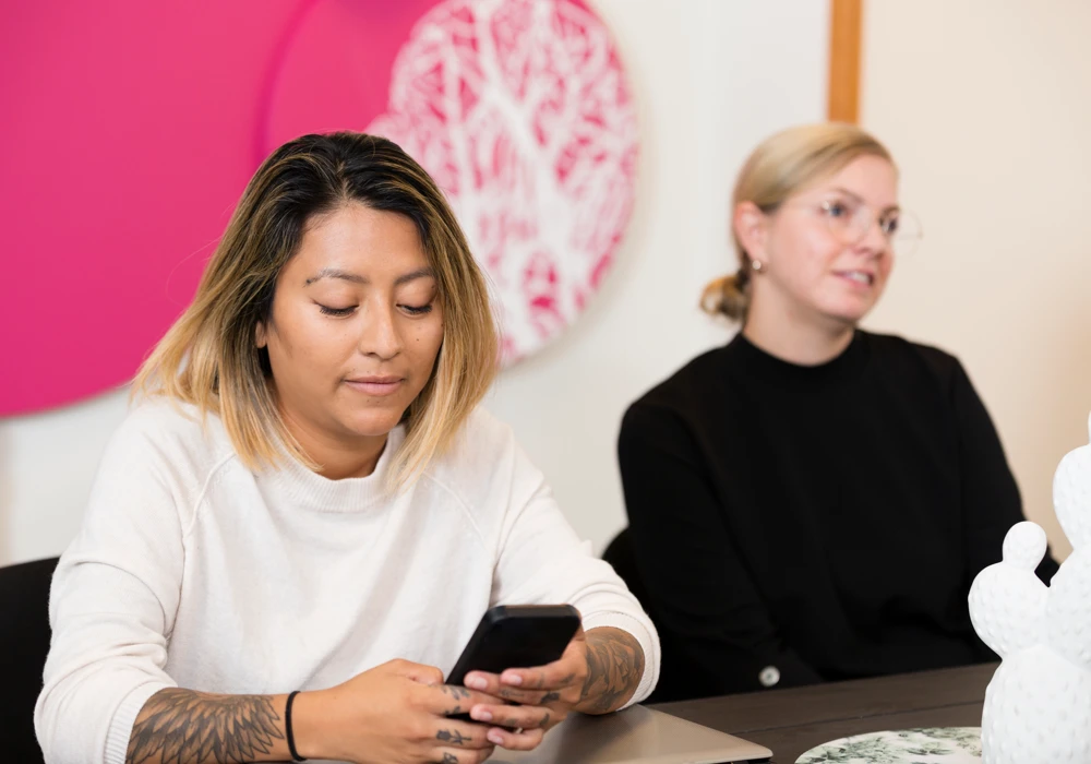 Två kvinnor sitter vid ett bord, och en tittar i sin mobiltelefon.
