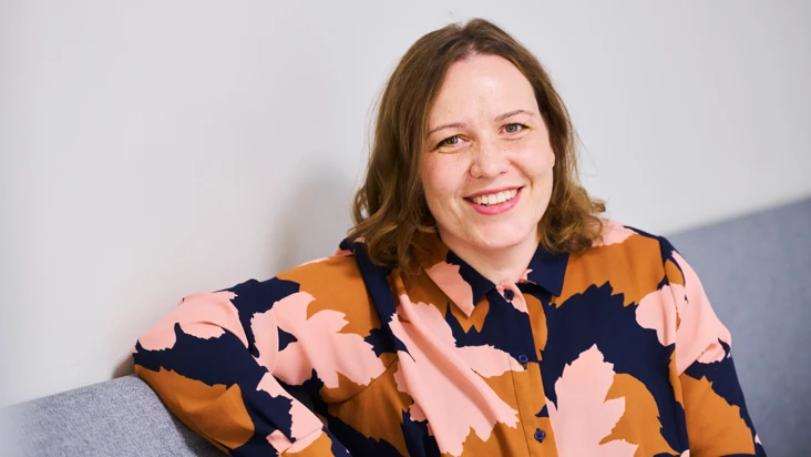 kvinna i blommig blus sitter i soffa och ler mot kameran.