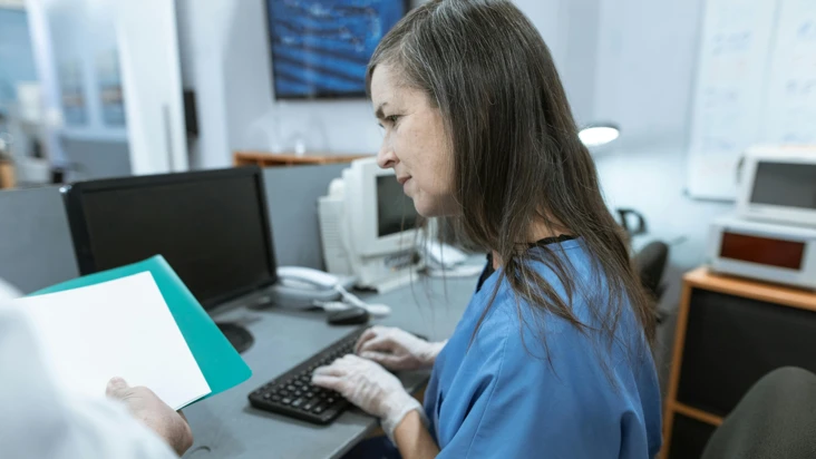 en kvinna i blå sjukhuskläder försöker skriva in något i datorn, som hon läser på ett papper bredvid.