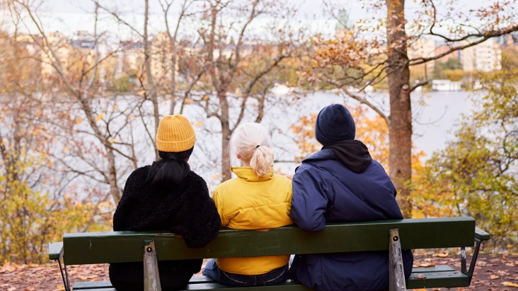 tre personer sitter nära varandra på en parksoffa i höstigt landskap