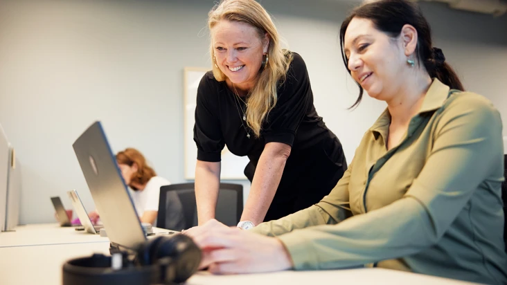 två kvinnor vid ett skrivbord i ett aktivitetsbaserat kontor. En står och en sitter och de tittar tillsammans på en dator.