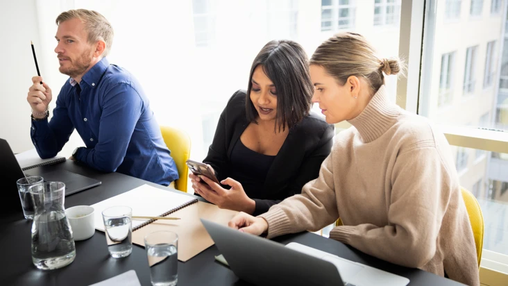 två kvinnor vid etts kjrivbord titta prå något i en mobil tillsammans och skrattar. En man sitter bredvid och ser allvarlig ut.