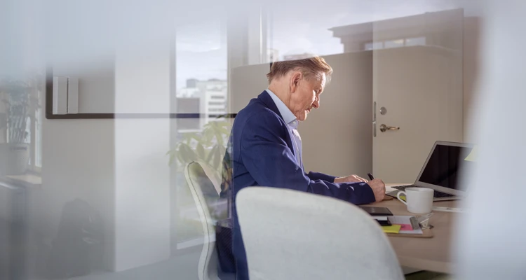 En äldre man i kostym jobbar ensam inne på ett kontor bakom en glasvägg.