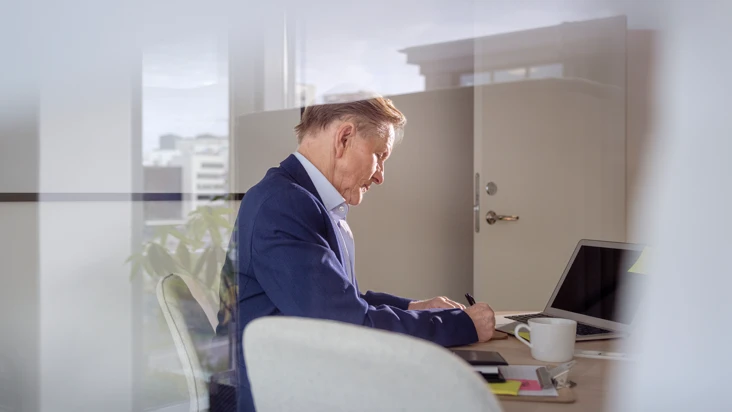 En man i bl&aring; kostym sitter bortv&auml;nd vid ett skrivbord och jobbar f&ouml;r sig sj&auml;lv.