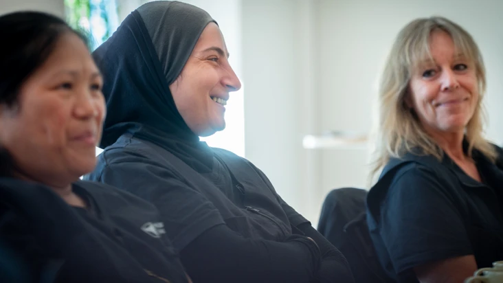 Tre kvinnor i olika &aring;ldrar sitter vid ett bord med solljus bakifr&aring;n och ler mot n&aring;gon utanf&ouml;r bilden.