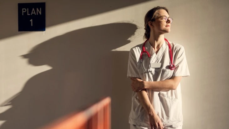 Kvinna i sjukhuskl&auml;der som st&aring;r mot en v&auml;gg och tittar mot solljuset.