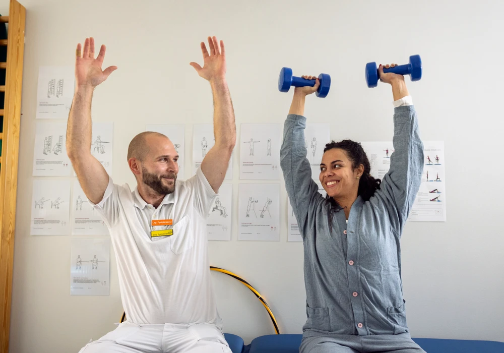 Patient tränar hos fysioterapeut. Hon håller hantlar ovanför huvudet.