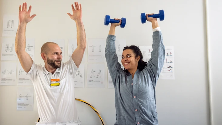 Kvinna och man tr&auml;nar med uppstr&auml;ckta armar i ljust tr&auml;ningsrum. Kvinnan h&aring;ller i ett par hantlar.