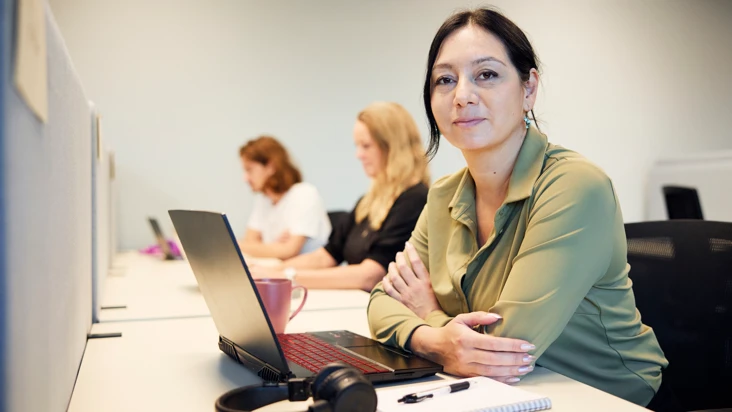 En kvinna i medel&aring;ldern med m&ouml;rkt h&aring;r och gr&ouml;n skjorta sitter vid en dator vid ett l&auml;ngre, delat skrivbord, och tittar in i kameran.