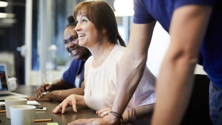 en glad kvinna sitter vid ett m&ouml;tesbord med andra omkring sig, som syns mer i bakgrunden.