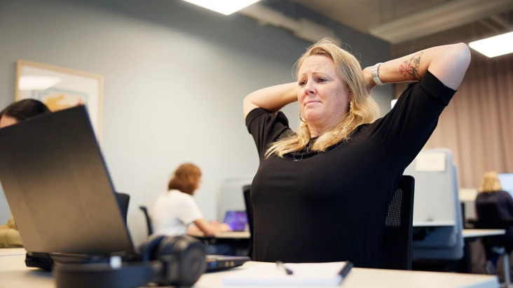 en kvinna som sitter p&aring; sin stol p&aring; ett kontor str&auml;cker p&aring; ryggen. I taket blinkar lamporna.