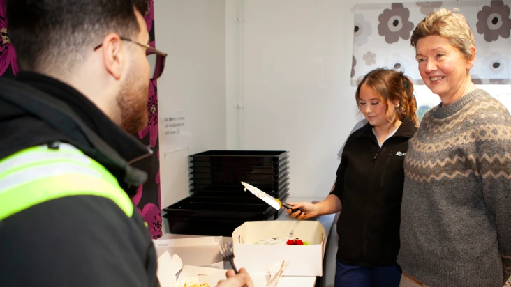 en man till v&auml;nster i arbetskl&auml;der med ryggen mot kameran, och en kvinna i m&ouml;nstrad ulltr&ouml;ja till h&ouml;ger. I mitten en ung kvinna som hugger in p&aring; en t&aring;rta med en br&ouml;dkniv.