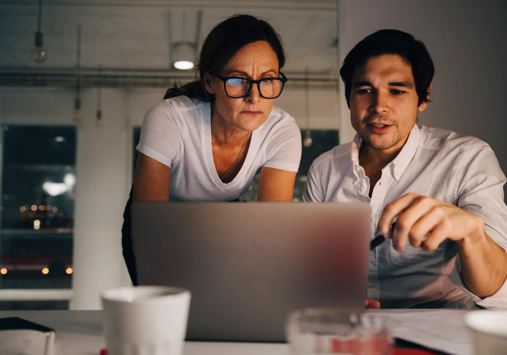 två personer tittar i en dator, i ett ödsligt kontorslandskap kvällstid.