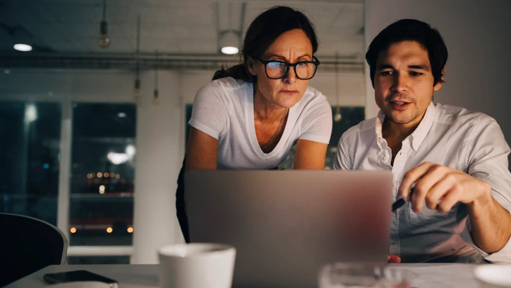 en man och en kvinna tittar i en dator tillsammans, i ett nedsl&auml;ckt kontorslandskap med kv&auml;llsm&ouml;rker utanf&ouml;r.