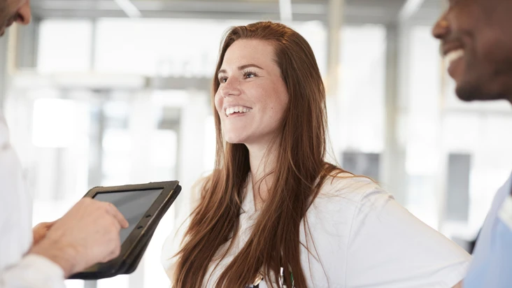 ung kvinna med l&aring;ngt brunt h&aring;r, i vit tr&ouml;ja, ler mot n&aring;gon till v&auml;nster som inte syns i bild. Till h&ouml;ger en man i profil.