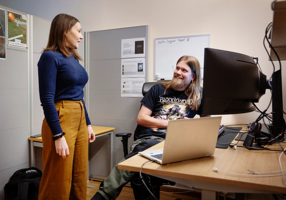 Kontorsrum med en man som sitter vid skrivbord med datorer, kvinna som st&aring;r. 