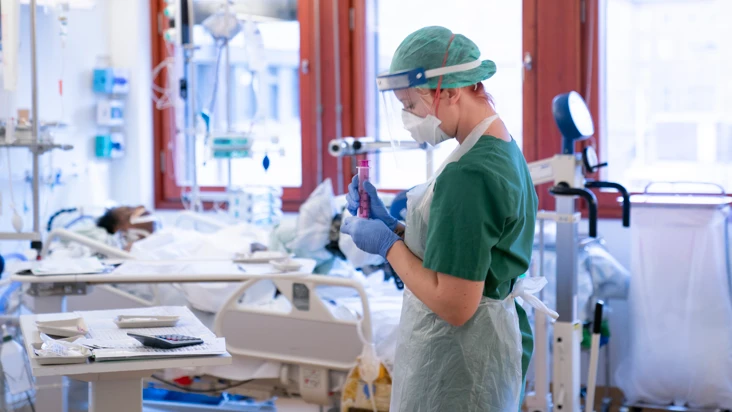 en sjuksk&ouml;terska iu skyddsutrustning i en sjukhussal. i bakgrunden en person som ligger i en s&auml;ng, uppkopplad till flera sladdar.