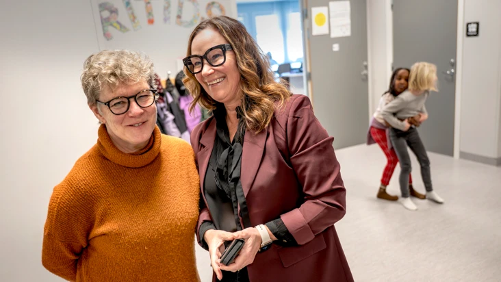 tv&aring; kvinnor i orange och lial kl&auml;der, st&aring;r i en hall med ett par barn bakom sig. P&aring; v&auml;ggen st&aring;r orden fritids.