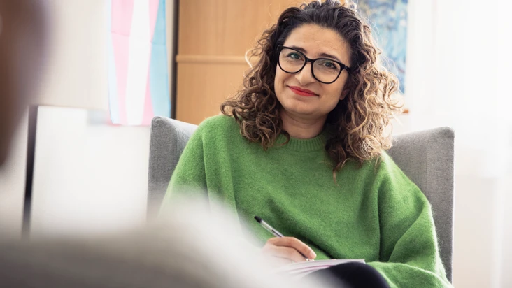 kvinna i gr&ouml;n tr&ouml;ja sitter i stol med penna och papper i handen. Hon ser p&aring; n&aring;gon mitt emot som inte syns i bild.