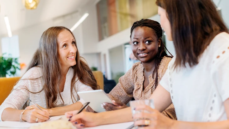 tre kvinnor talar engagerat med varandra vid ett bord. I bakgrunden bokhyllor.