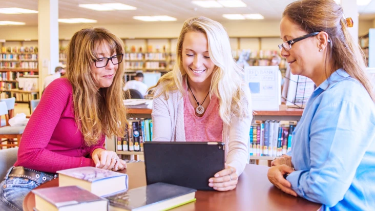 Tre kvinnor st&aring;r samlade vid ett st&aring;bord i ett bibliotek.