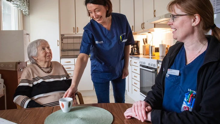&auml;ldre dam i rullstol sitter vid k&ouml;ksbord och tittar upp p&aring; tv&aring; glada undersk&ouml;terskor som serverar kaffe vid bordet.
