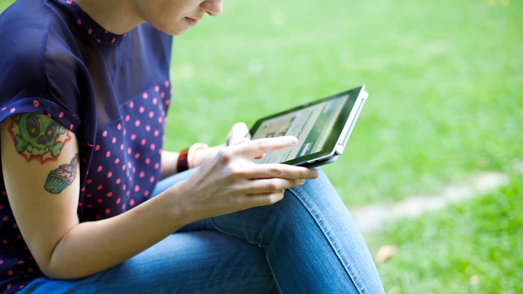 kvinna i bl&aring; blus sitter med en Ipad utomhus vid gr&auml;smatta.