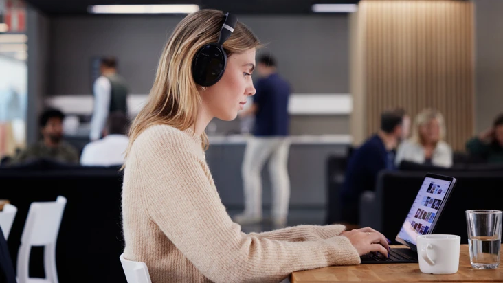 ung kvinna i kontorslandskap sitter och skriver p&aring; en dator, med ett par stora h&ouml;rlurar p&aring; sig.