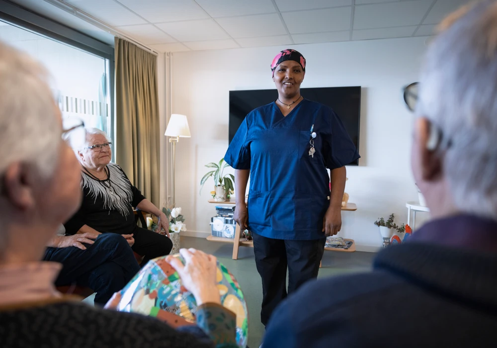 Undersköterska i äldreomsorgen står och ler framför några sittande äldre personer.