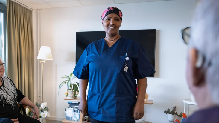 kvinnlig undersk&ouml;terska i bl&aring; arbetskl&auml;der st&aring;r framf&ouml;r en grupp sittande &auml;ldre i ett vardagsrum p&aring; ett &auml;ldreboende.