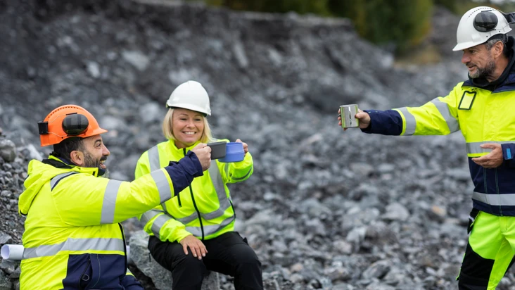 tv&aring; kvinnor och en man i gula skyddskl&auml;der sitter i en grusig backe och sk&aring;lar glatt med sina termosmuggar.