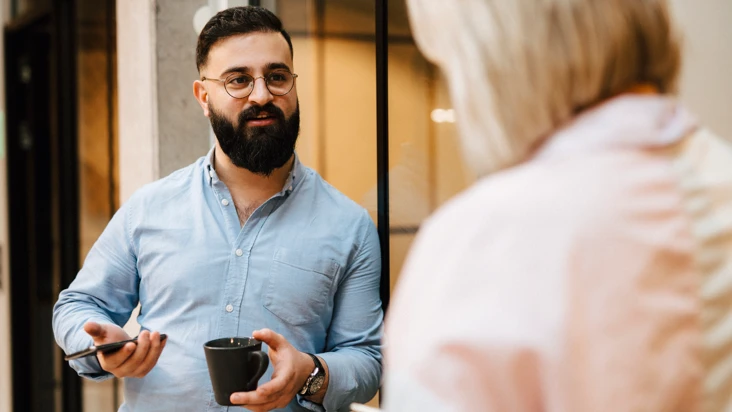Man i bl&aring; skjorta v&auml;nder sig mot kvinna, utomhus, med mobil i ena handen, och en kopp kaffe i den andra handen.