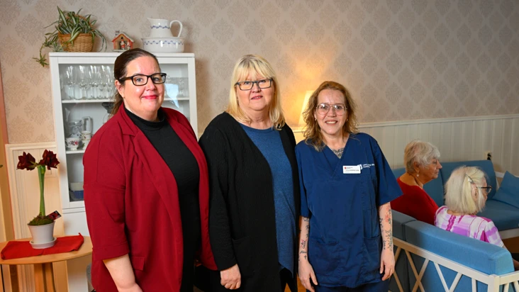 Tv&aring; kvinnor i kofta och tr&ouml;ja, samt en kvinna i bl&aring; arbetskl&auml;der f&ouml;r undersk&ouml;terska. De st&aring;r i ett trivsamt rum. Bakom dem tv&aring; &auml;ldre kvinnor i en soffa med ryggen mot fotografen.