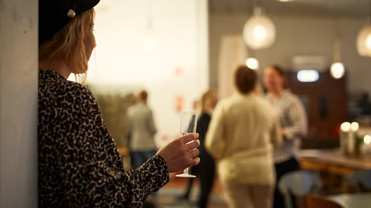 kvinna med ryggen mot en v&auml;gg och ett glas i handen ser bort mot ett par andra personer som st&aring;r och pratar med varandra med varsitt glas i h&auml;nderna.