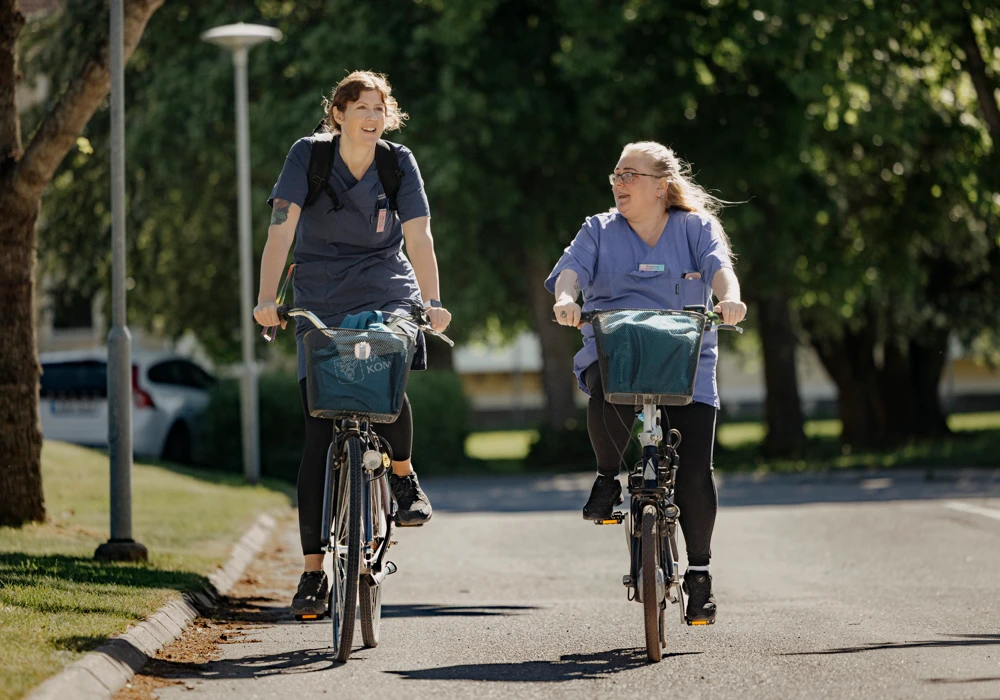 tv&aring; kvinnliga undersk&ouml;terskor i bl&aring; arbetskl&auml;der cyklar p&aring; mindre v&auml;g med gr&ouml;nska i bakgrunden.