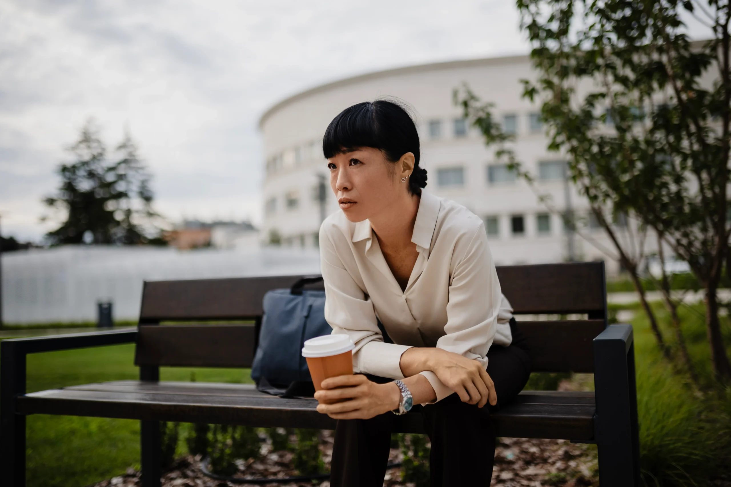 En kvinna sitter utomhus på en bänk och reflekterar med en kopp kaffe i handen.