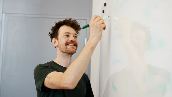 Leende man med mustasch och ikl&auml;dd t-shirt skriver n&aring;got p&aring; en vit tavla.