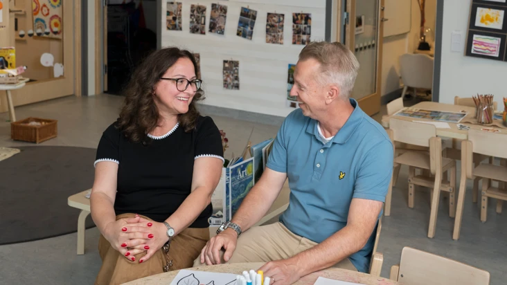 en kvinna och en man sitter och samtalar p&aring; l&aring;ga barnm&ouml;bler i en f&ouml;rskolemilj&ouml;.