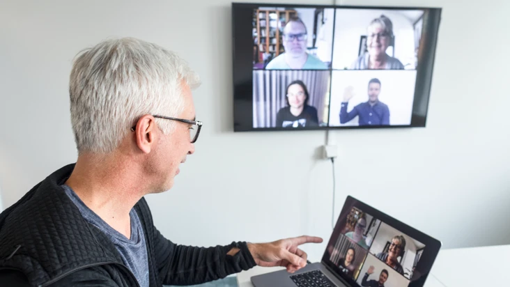 En man sitter vid ett bord och tittar på en dator där det pågår ett digitalt möte. På väggen en skärm med samma digitala möte.